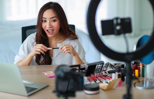 Селективный фокус на камеру, молодая и красивая азиатская девушка показывает макияж, как использовать помаду, на камеру с улыбкой и счастливой во время трансляции видеозаписи о косметическом содержании и обзоре.