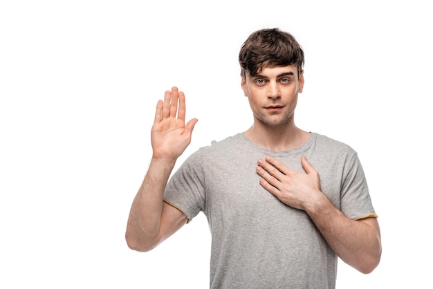 Photo serious young man looking at camera while showing swear gesture isolated on white