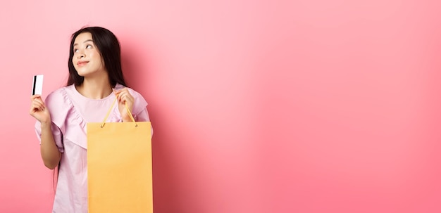 Фото Шоппинг мечтательная азиатская девушка думает о покупке чего-то, держа в руках сумку и пластиковую кредитную карту