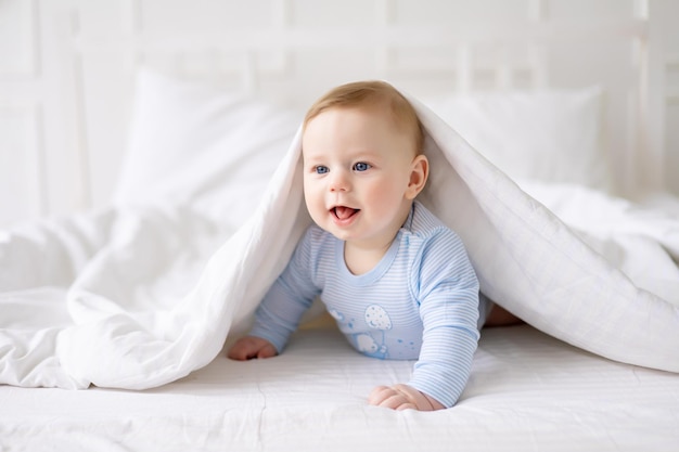 Photo a small healthy baby is lying at home under a blanket on white bed linen in a blue bodysuit happy baby lying on his stomach textiles and bedding for children
