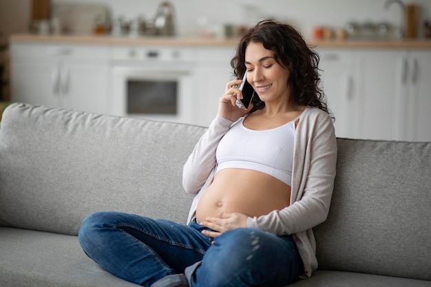 Smiling pregnant woman talking on cellphone and caressing her belly while relaxing on couch at home