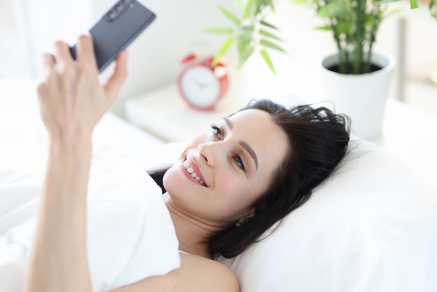 Улыбающаяся женщина лежит в постели и смотрит в смартфон