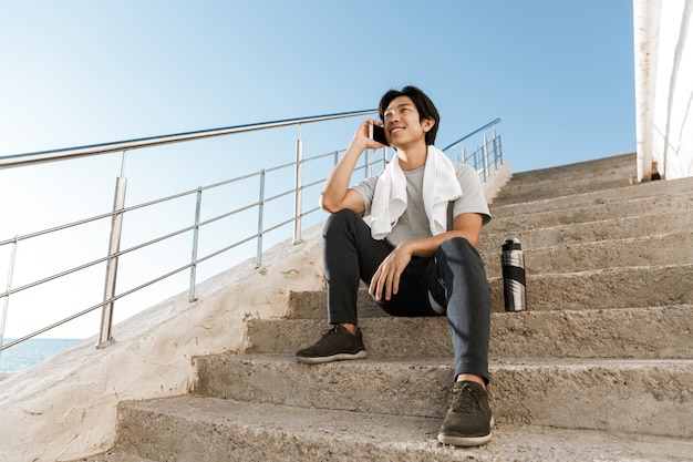 사진 해변에서 계단에 앉아 젊은 아시아 운동가 웃고, 휴대 전화로 이야기
