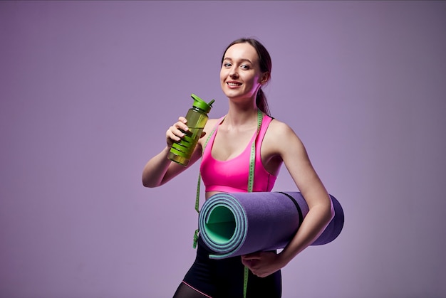 Фото Спортивная красивая женщина в спортивной одежде, держащая бутылку воды и фитнес-циновку, изолированную на фиолетовом. иди в тренажерный зал.