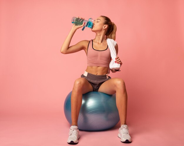 Фото Спортивная женщина, сидящая на фитболе с питьевой водой на розовом фоне