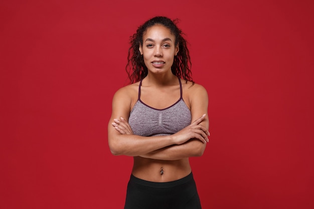 Фото Сильная молодая афро-американская спортивная женщина фитнеса в спортивной одежде, позирующая тренировка, изолированная на портрете красной стены. спорт осуществляет концепцию здорового образа жизни. держась за руки скрещенными.