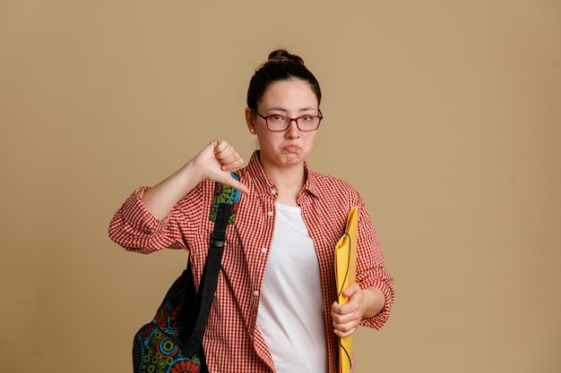 Student young woman in casual clothes wearing glasses with backpack holding folder looking at camera displeased and offended showing thumb down standing over brown background