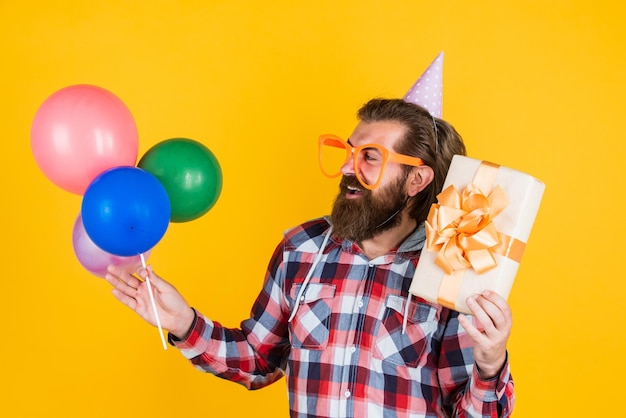 Стильный бородатый парень с модной прической носит клетчатую рубашку, держит воздушные шары и подарит коробку на день рождения