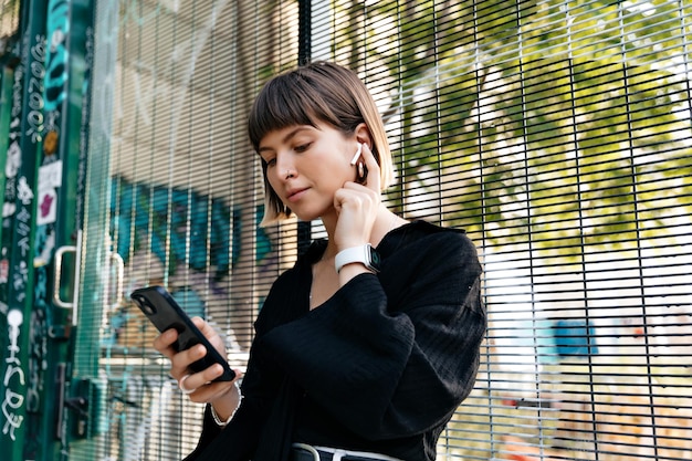 Stylish pretty girl with light short hairstyle wearing black blouse listening music in wireless headphone and holding smartphone in sunlight Recreation outdoor in summer evening