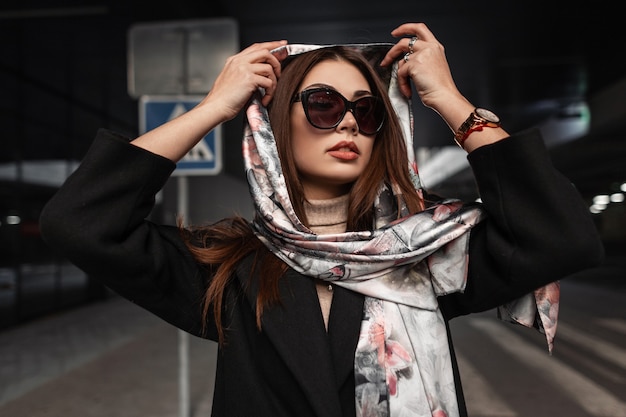 Photo stylish young european woman with sexy beautiful lips in trendy sunglasses in coat puts elegant silk scarf on head in the city. portrait business fashion model girl. vintage beauty lady on street.