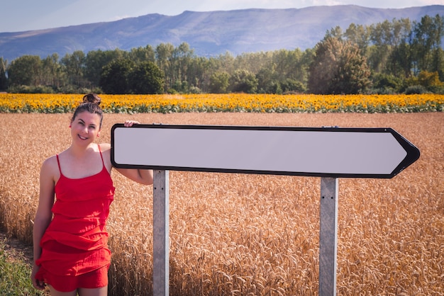 Gratis foto betoverend shot van een aantrekkelijke vrouw in een rode jurk poserend in een tarweveld met verkeersbord