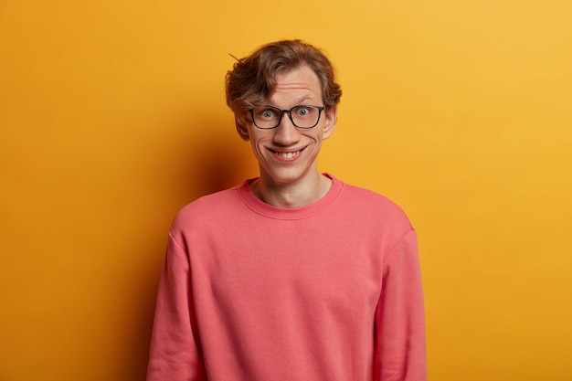 Gratis foto geïsoleerd beeld van positieve hipster-man heeft een gelukkige reactie op recent nieuws, in een goed humeur, kijkt verrassend door een bril, draagt een casual roze trui, geïsoleerd op een gele muur. echte menselijke emoties