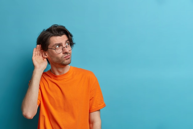 Gratis foto foto van nieuwsgierige man houdt de hand in de buurt van het oor en luistert naar privé-informatie, probeert roddelen af te luisteren, heeft een geïntrigeerde uitdrukking, draagt een ronde bril en een oranje t-shirt, kopie ruimte op blauwe muur