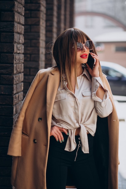 Gratis foto jonge vrouw in beige jas die telefoon op straat gebruikt