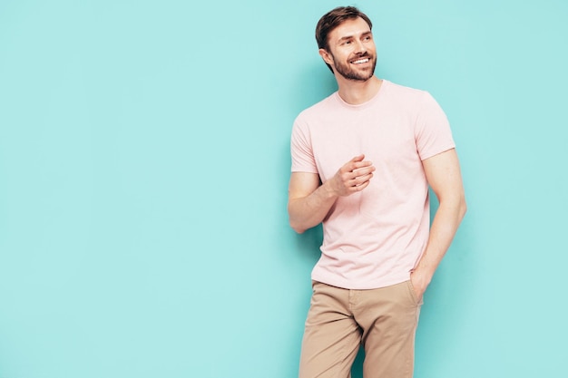 Gratis foto portret van knappe lachende stijlvolle hipster lamberseksueel model sexy man gekleed in roze tshirt en broek mode man geïsoleerd op blauwe muur in studio
