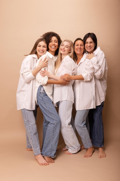 Gratis foto volledige lengte gelukkige dames van verschillende leeftijden in witte overhemden en spijkerbroeken die naar de camera glimlachen tegen een beige achtergrond