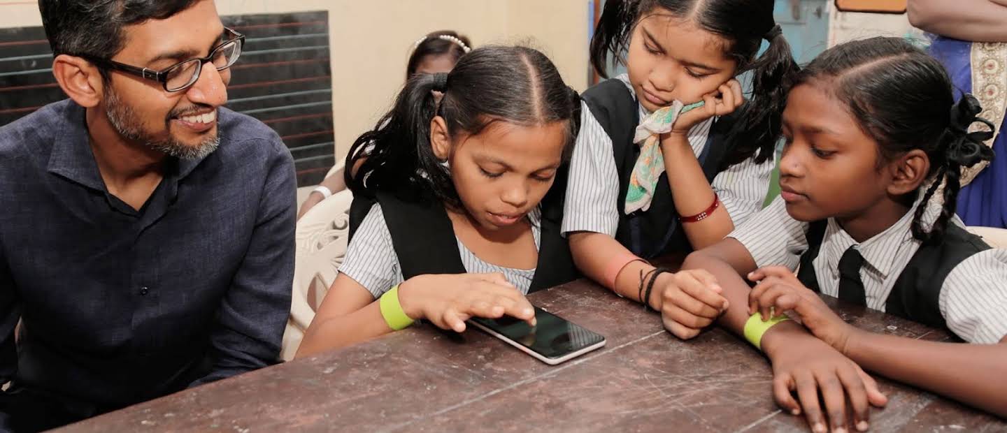 Sundar Pichai sammen med tre skolejenter i uniform – alle konsentrerer seg om en smarttelefon