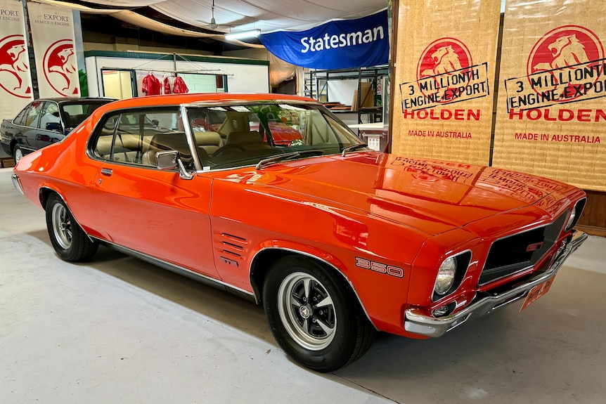 An orange-red 1972 Holden Monaro in front of several Holden signs. 