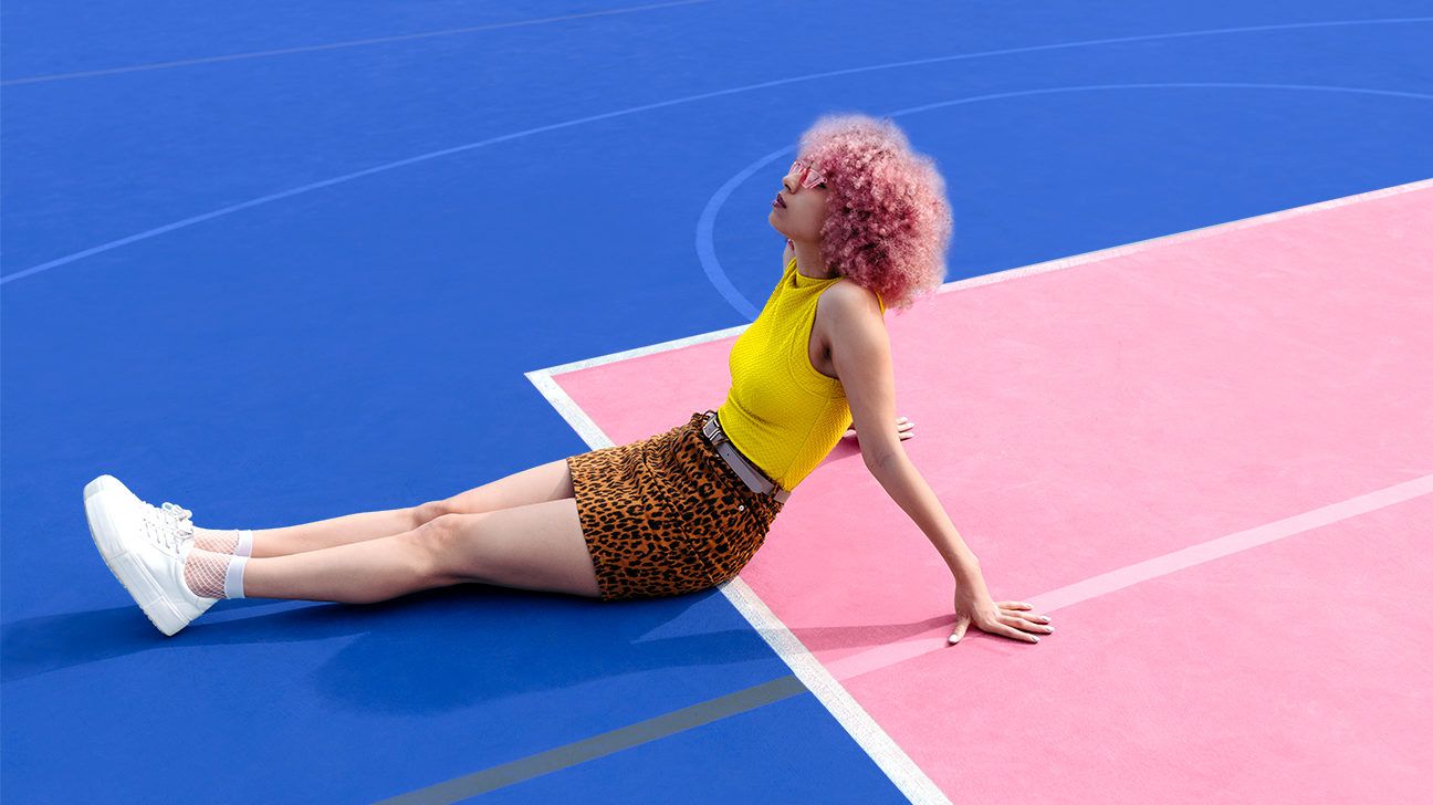 Stylish girl with a pink afro, leopard skirt, and yellow halter top, daydreams while lounging on a pink and blue tennis court. 