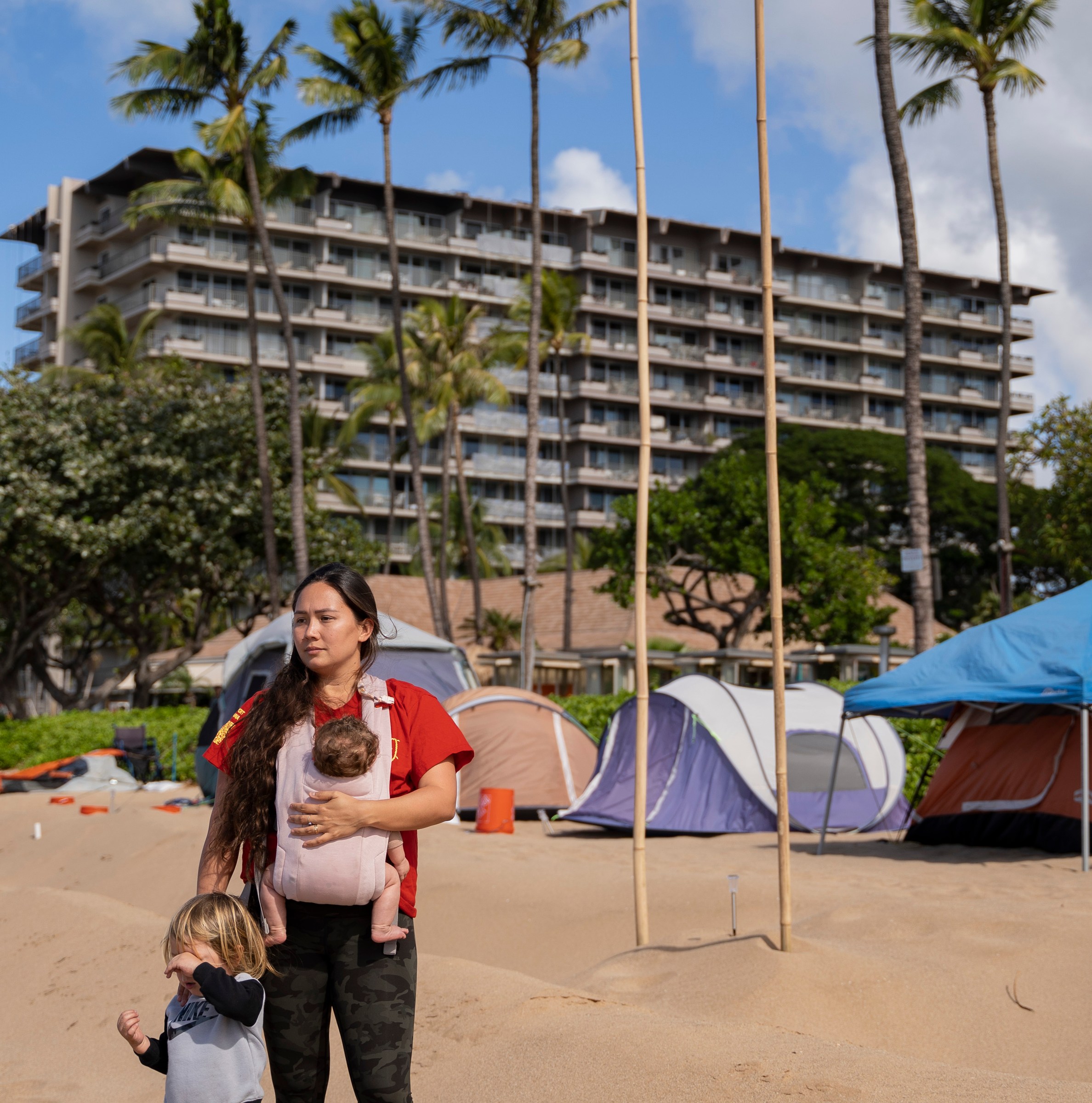 A woman with two young children stands on a beach in front of a multistory hotel, palm trees, and tents.