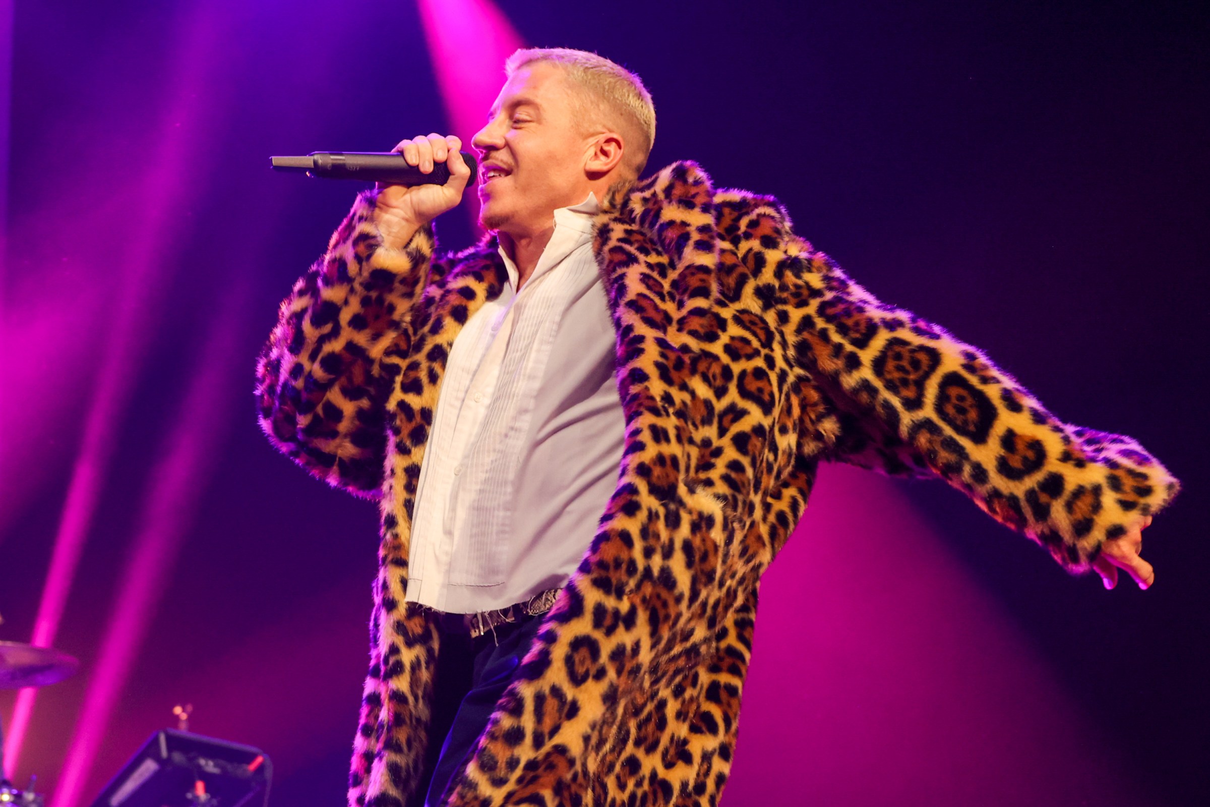 Macklemore, wearing a leopard print coat and lit by purple and pink stage lights, sings into a handheld microphone.