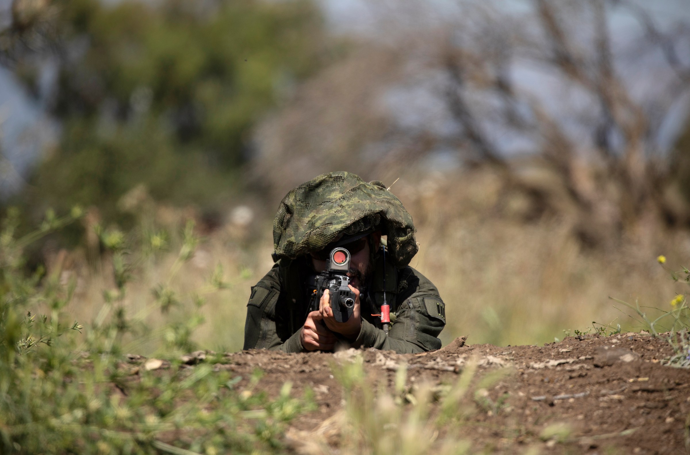 An Israeli soldier lying on the ground amid shrubs points a rifle directly at the camera.