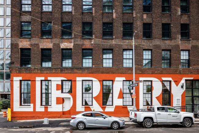 An exterior shot of Adams Street Library. The building is painted orange with the word "Library" printed in white letters across the first floor of the building.