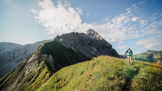 Wandernde Person auf einem grün bewachsenen Bergkamm, im Hintergrund blauer Himmel mit weißen Wolken.