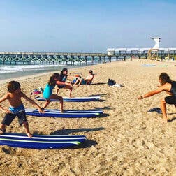 On a sandy beach, three children practice surfing positions on striped surfboards that are placed on the sand, as their teacher, a man with shaggy hair and wearing black swimming gear, instructs them. In the background, beneath a blue sky, a long pier juts into the ocean.
