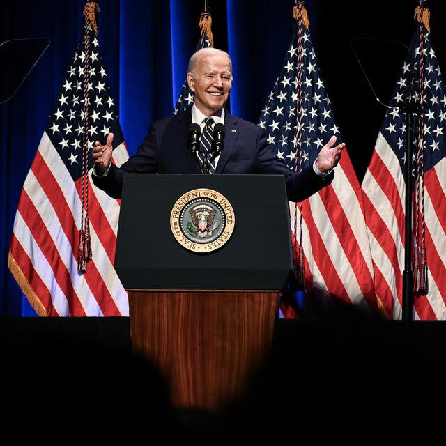 President Biden standing and smiling with his hands raised at a lectern in front of American flags.