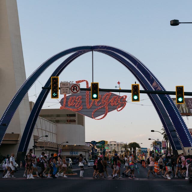 A neon Las Vegas sign has two blue arches and the words “Las Vegas” in script. Underneath, pedestrians walk in a crosswalk.