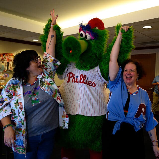 The Phillies green mascot, Philly Phanatic, is flanked by two smiling brides, casually dressed. Each is holding one of his arms up in the air. 