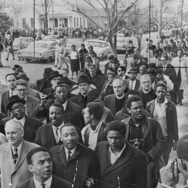 A black and white photograph of a protest march from 1965. Leading the crowd is Rev. Dr. Martin Luther King. In the background are police officers and cars. 