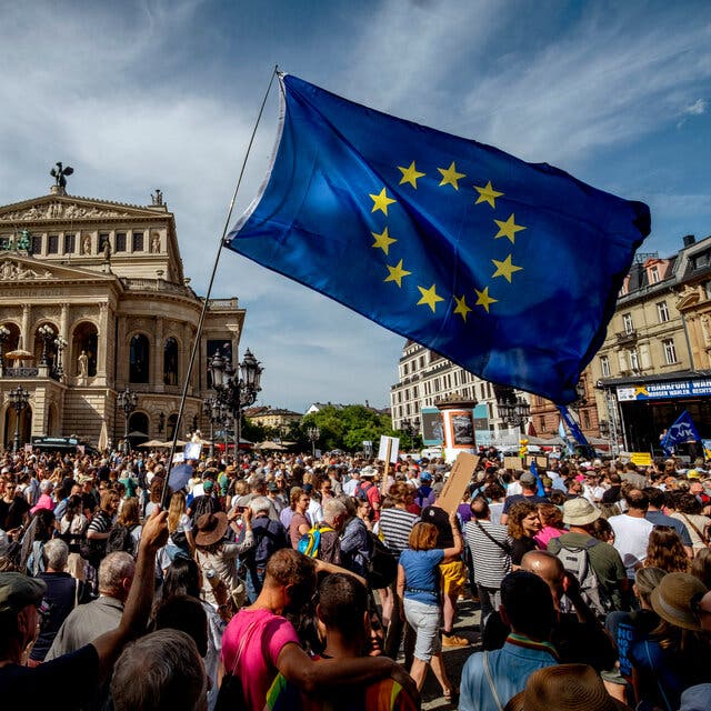 A large crowd in a plaza with a grand Renaissance-style building, Frankfurt’s Old Opera House, in the background. A blue European Union flag waves in the foreground.