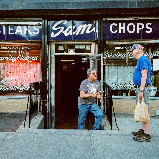 Louis Migliaccio, wearing jeans and a T-shirt, stands on the steps leading down to his restaurant, Sam’s, as he talks to a neighbor standing on the sidewalk above.